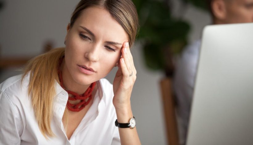 A woman in a white shirt looking stressed while sitting at her desk with a computer, touching her temple.