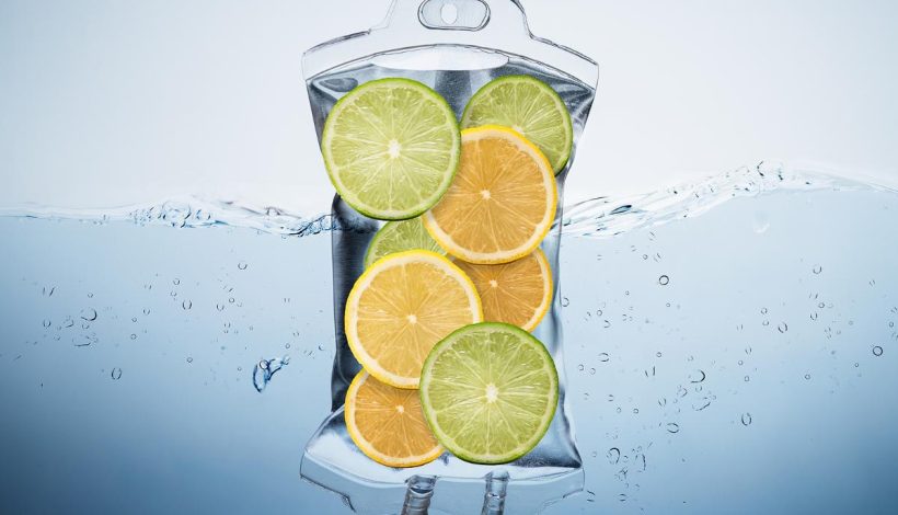 Transparent water pitcher filled with orange and lime slices, partially submerged in water with bubbles around it.