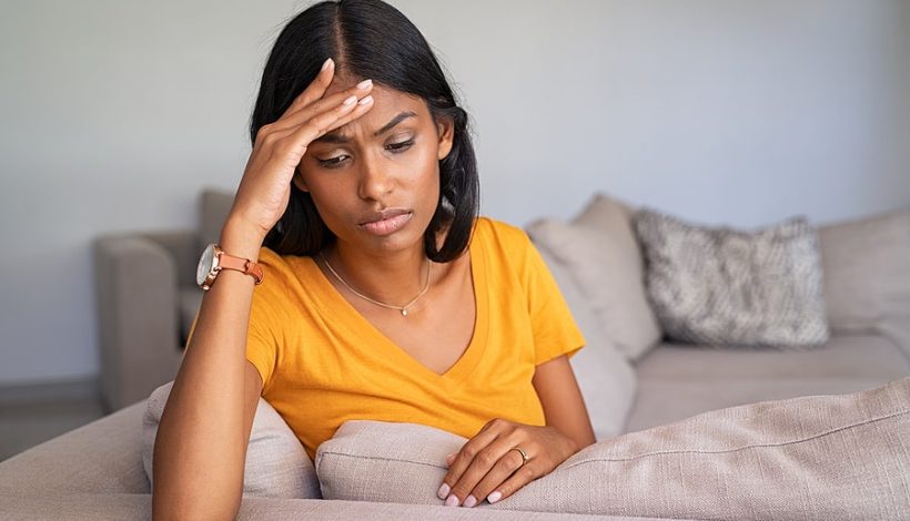 A woman sitting on a couch looking stressed with her hand on her forehead.
