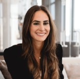 A woman with long brown hair, wearing a black top, sits indoors on a sofa, smiling at the camera.