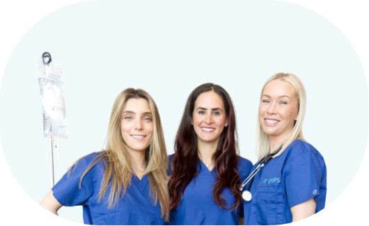 Three female healthcare professionals in blue scrubs standing together, smiling, with an iv stand in the background.