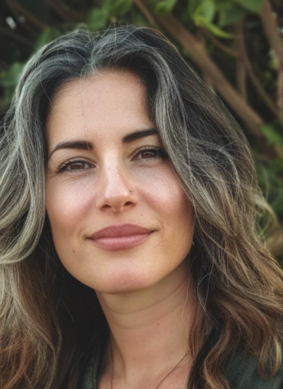 A woman with long, wavy, brown hair with gray streaks, and a light complexion, smiles softly at the camera. The background features green foliage.
