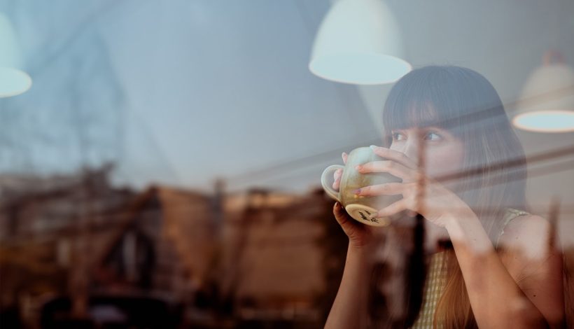 A woman drinking coffee at a cafe table, viewed through a window with reflections. she has bangs and is holding a large cup with both hands.