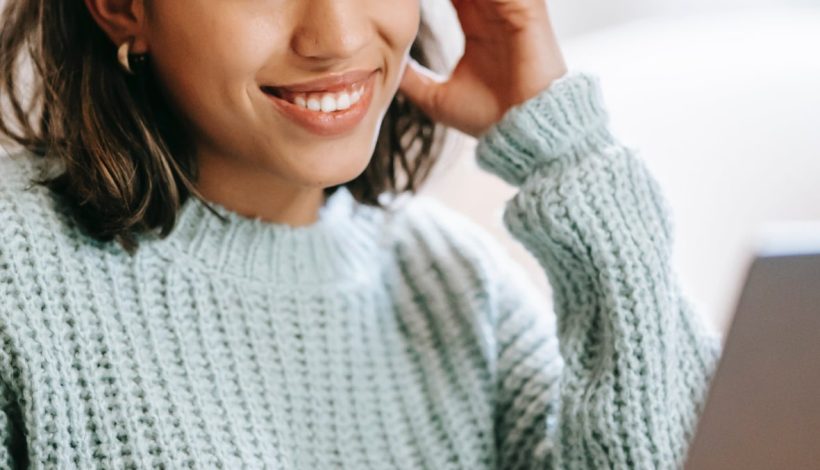 Young woman smiling and looking at her laptop screen, wearing a teal sweater, with her hand resting on her head.