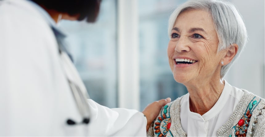 Elderly white woman smiling at a doctor, who is placing a comforting hand on her shoulder in a bright medical office.