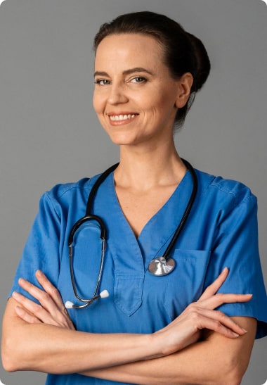 A confident female healthcare professional in blue scrubs with folded arms, wearing a stethoscope, smiling at the camera on a gray background.