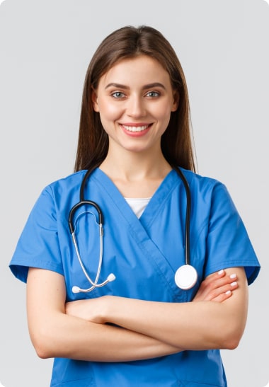 A young female nurse in blue scrubs with a stethoscope around her neck, smiling and standing with her arms crossed.