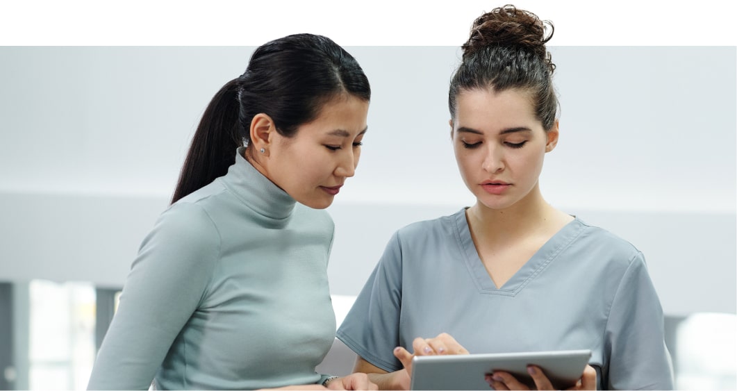Two women, one in a turquoise turtleneck and the other in a blue medical scrub, looking at a tablet together in a clinical setting.