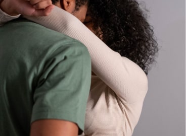 A person with curly hair sneezing or coughing into their elbow, wearing a green shirt and a beige long-sleeve undershirt.