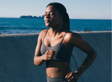 Young woman jogging by the sea on a sunny day, wearing a sports bra and leggings, with a focused expression.