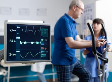 Senior man walking on treadmill under supervision of a female nurse with a monitor displaying vital signs in the background.