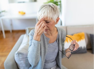 Senior woman with short white hair sitting on a couch, looking tired or distressed, holding her glasses in one hand and touching her forehead with the other.