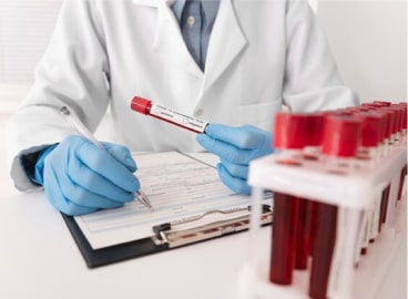 A medical professional in a lab coat and gloves examines a blood sample vial while writing notes, with a rack of other samples nearby.