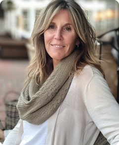 A middle-aged woman with shoulder-length blonde hair, wearing a beige scarf and white top, smiles gently while sitting outdoors.