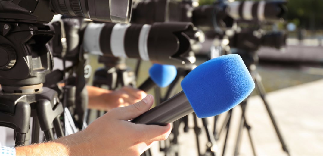 A row of video cameras with blue microphone covers, focused on an event, with a person's hand adjusting one of the microphones.