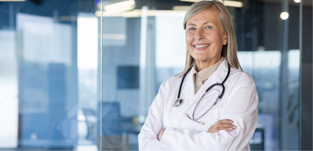 A mature female doctor with a stethoscope around her neck, smiling and standing confidently in a modern hospital environment.