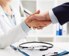 A doctor and a patient shaking hands over a desk with a stethoscope and medical documents visible.