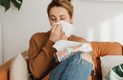A woman sitting on a couch, blowing her nose with a tissue, holding a floral box of tissues, wearing a brown sweater and ripped jeans.
