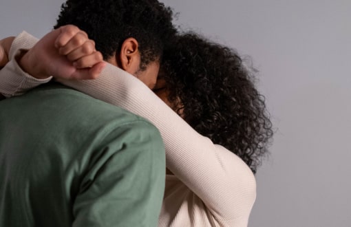 Two individuals embracing, one with curly hair and the other with short hair, both wearing long sleeves, against a plain background.