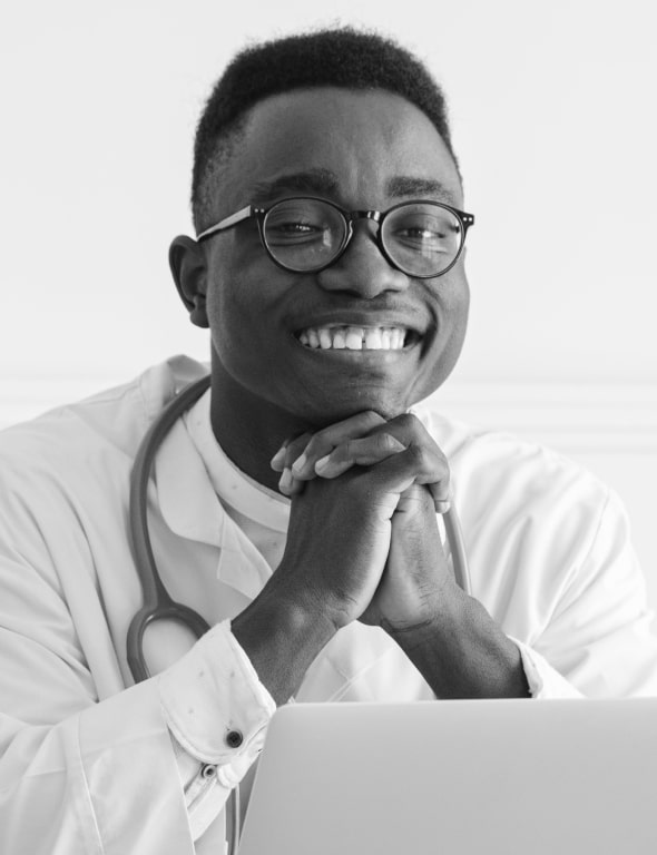 A smiling black male doctor with glasses sitting at a desk, resting his chin on his hands in front of a laptop, in a white coat with a stethoscope around his neck.