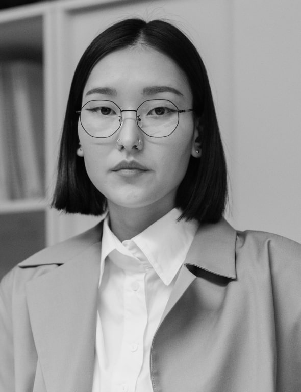 Black and white portrait of a woman with glasses, wearing a white shirt and grey blazer, looking directly at the camera.