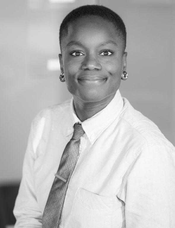 Black-and-white portrait of a smiling black woman wearing a white shirt and tie, earrings visible, looking at the camera.