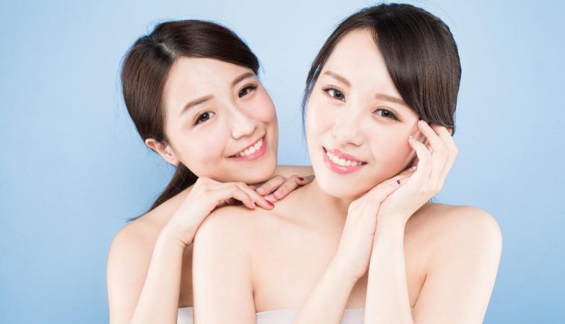 Two smiling women with bare shoulders posing close together against a light blue background.