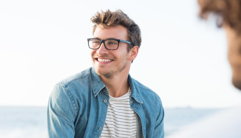 Smiling man with glasses wearing a denim jacket and striped shirt, looking at someone off-camera by the sea.