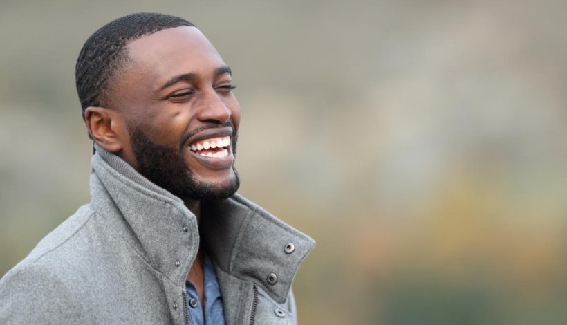A joyful black man with a beard, wearing a gray jacket, laughing outdoors with a blurred natural background.