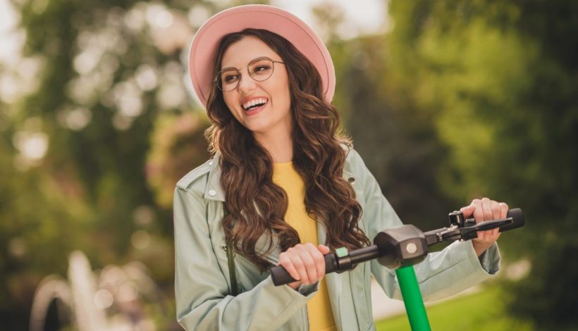 A joyful woman in a pink hat and glasses rides an electric scooter in a sunny park.