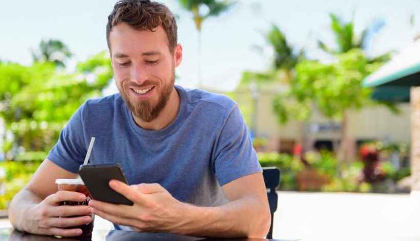 A man smiling as he uses a smartphone at a cafe table outdoors, holding a drink with a straw.