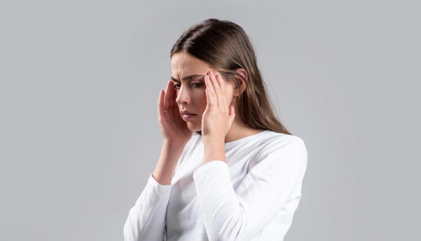 A woman in a white shirt touching her temples, looking stressed or in pain, against a gray background.