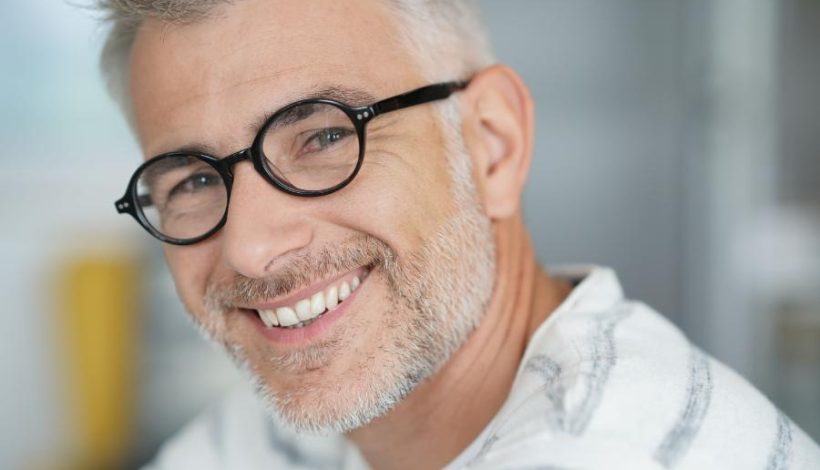 A man with gray hair and beard, wearing round glasses and a white shirt with gray stripes, smiles at the camera.
