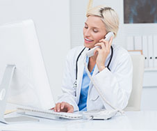 A doctor in a white coat is sitting at a desk, talking on a telephone, and working on a computer.