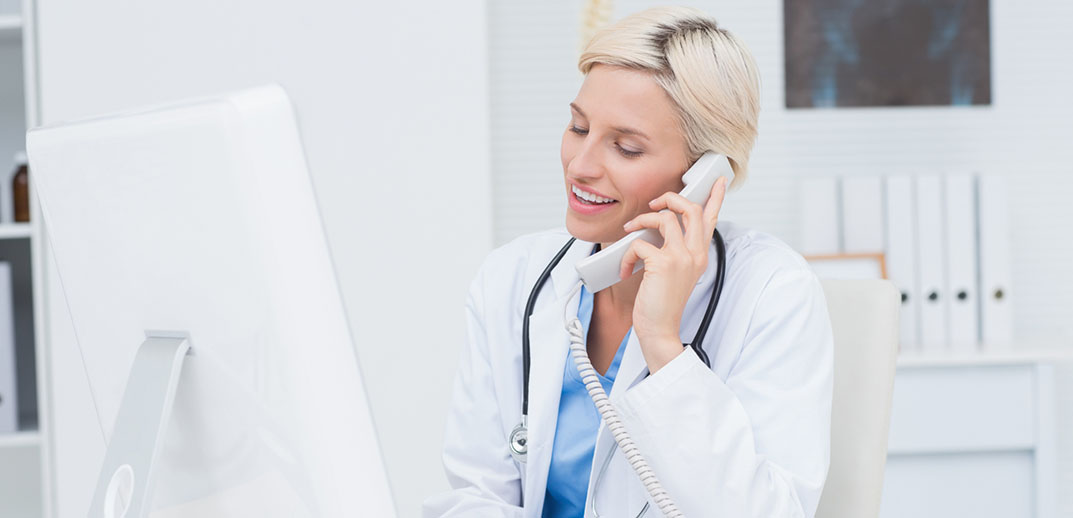 A doctor in a white coat and stethoscope sits at a desk, smiling while talking on the phone.