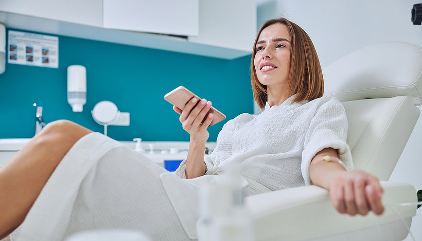 A woman in a white robe sits in a medical chair, holding a smartphone in one hand with a medical bandage on her other arm. She appears to be in a clinic or medical facility.