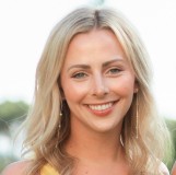 A woman with long blonde hair smiles at the camera, wearing a yellow top and dangling earrings, with a blurred outdoor background.