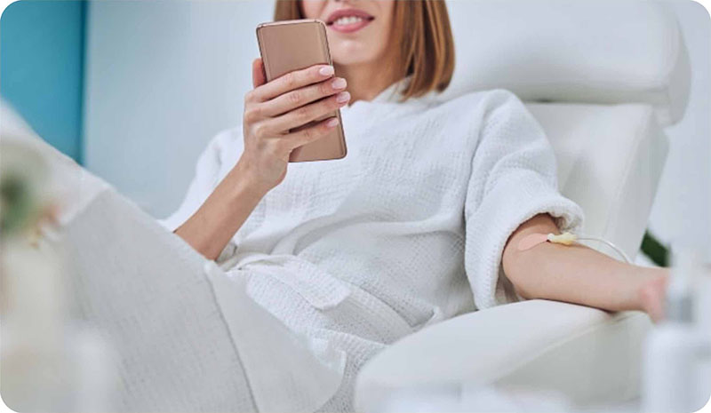 A person in a white blanket holds a smartphone, partially covering their face, while resting on a sofa.