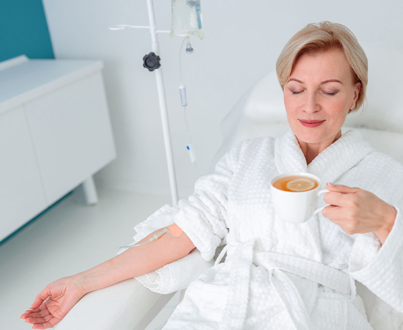 A woman in a white robe enjoys a cup of tea while receiving an iv treatment in a modern clinic room.