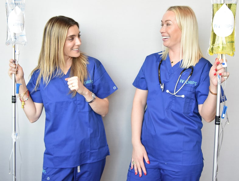 Two women in blue medical scrubs stand holding IV bags, smiling and looking at each other against a plain background.